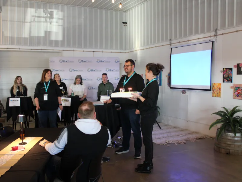 Group of people in a casual indoor event with a woman holding a cake with lit candles and others seated or standing.