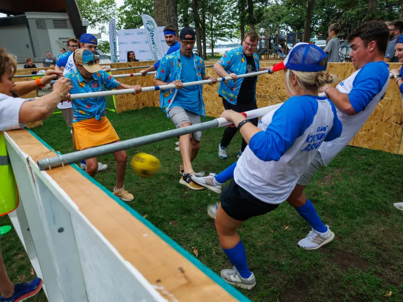 People playing human foosball outdoors in a wooden arena wearing blue and white jerseys on grass.