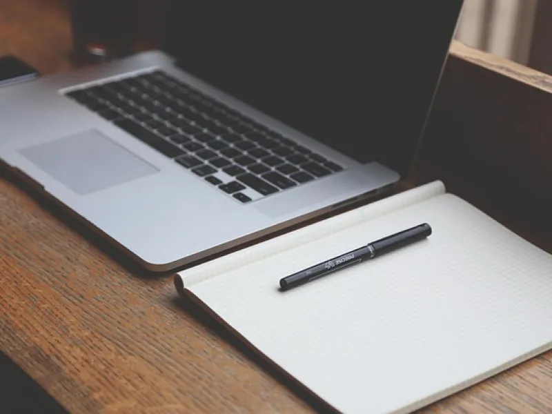 Open laptop beside blank grid notebook with pen on wooden desk workspace setup.