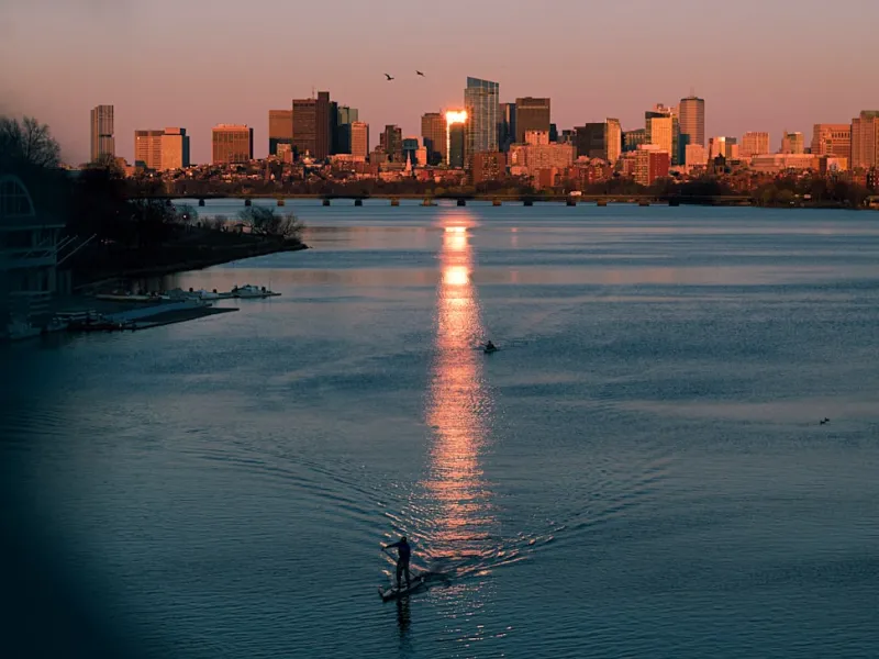 Paddleboarder on river at sunset with city skyline and reflective water under warm light.