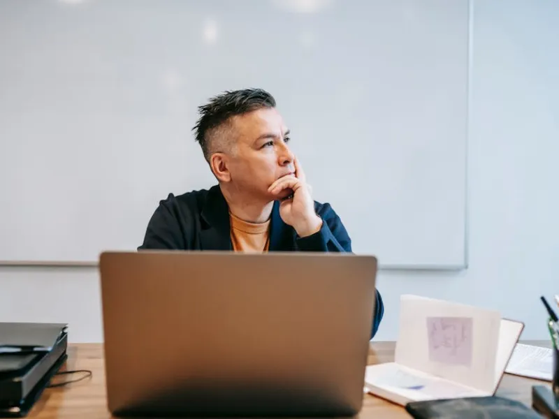 Man in casual jacket sitting at desk with laptop, looking thoughtfully to side in bright office space
