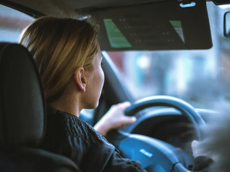 Woman with blonde hair driving a car, seen from the back seat inside the vehicle during daytime.