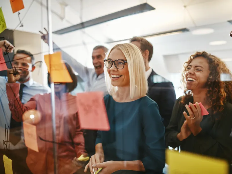 Diverse team collaborating happily around glass wall with colorful sticky notes and diagrams in modern office.