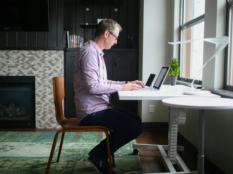 Man working on laptop at a white desk near window in a modern living room with fireplace and green rug