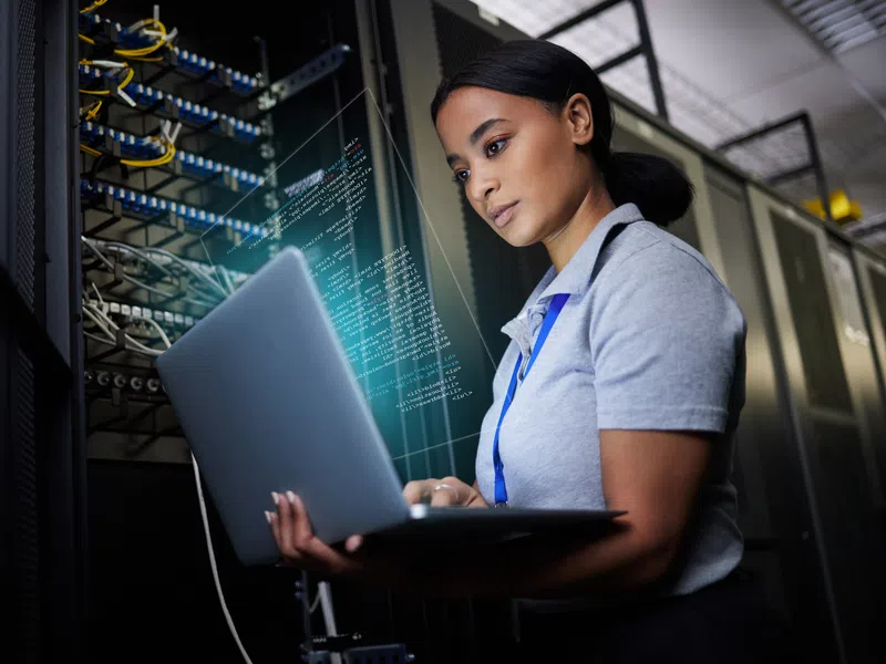 Young female IT technician working on laptop in server room with digital code overlay.