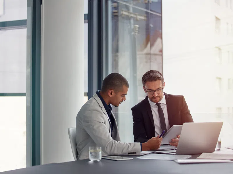 Two businessmen discussing documents at a desk with laptop in a modern office with large windows.