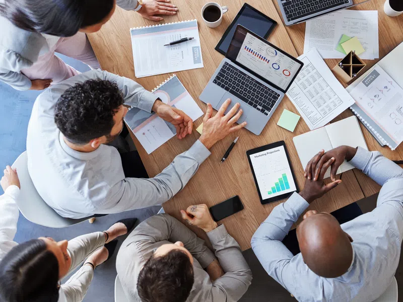 Business team collaborating around a table with laptops, charts, documents, and coffee cups in a meeting.