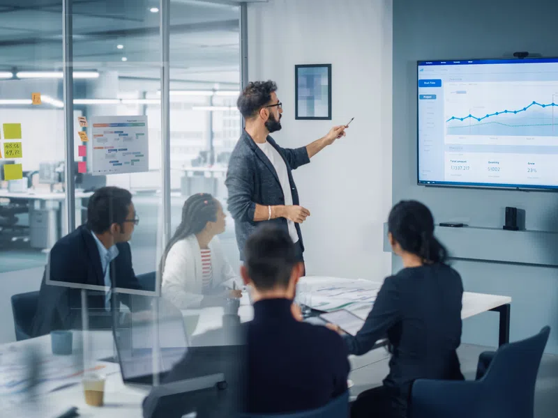 Business team in meeting room watching man present data analytics on large screen with graphs and charts
