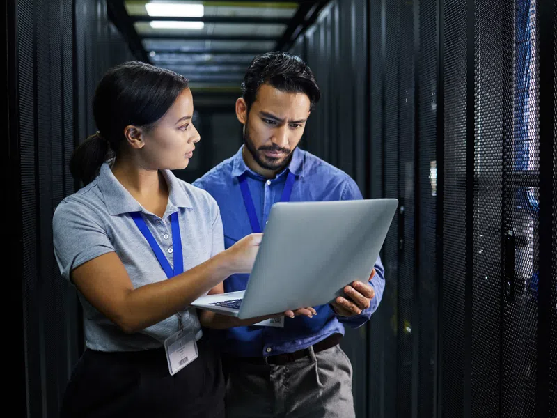 Two IT professionals examining data on a laptop inside a modern data center server room.