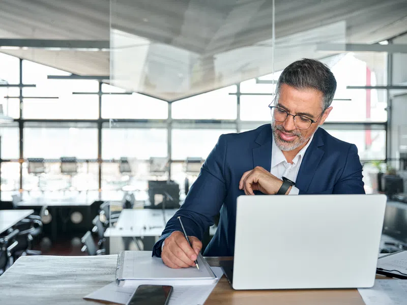 Businessman in a blue suit working on laptop while taking notes in a bright modern office.