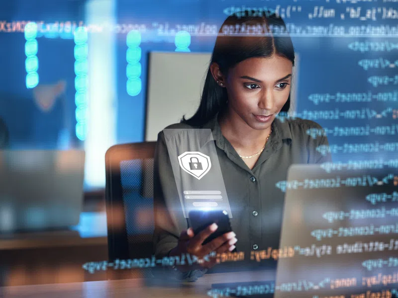 Focused woman using smartphone with digital security icons and coding overlays in modern tech workspace.