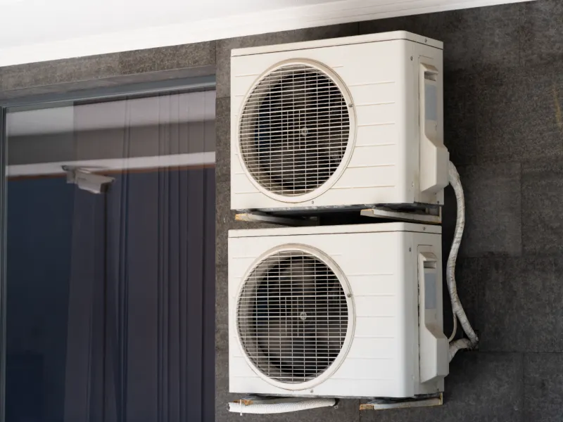 Two white outdoor air conditioning units mounted vertically on a dark exterior wall near a window.