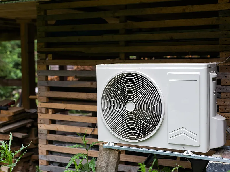 Outdoor air conditioning unit mounted on a wooden structure with stacked firewood in the background.