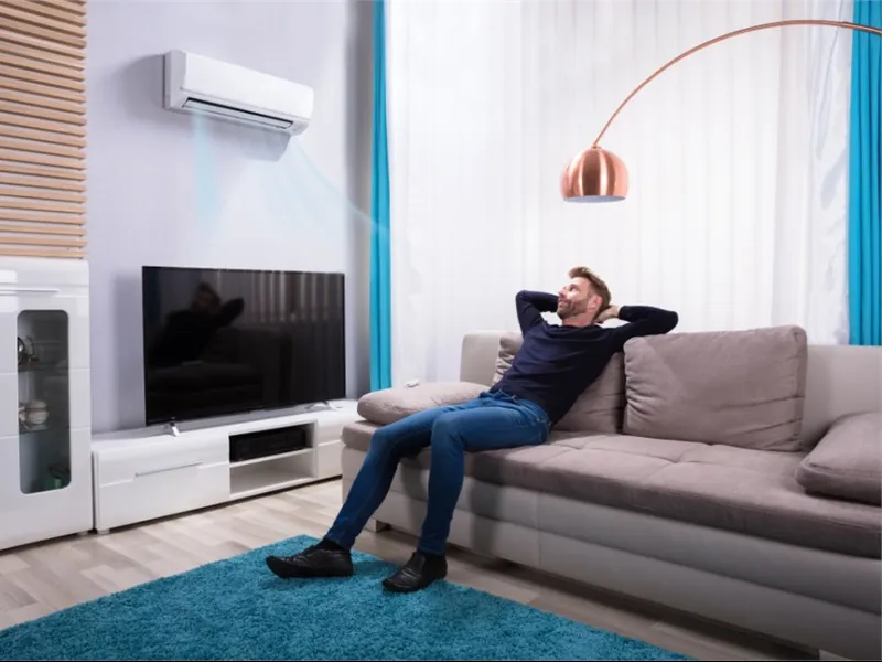 Man sitting relaxed on sofa in living room with air conditioner blowing cool air and modern decor.