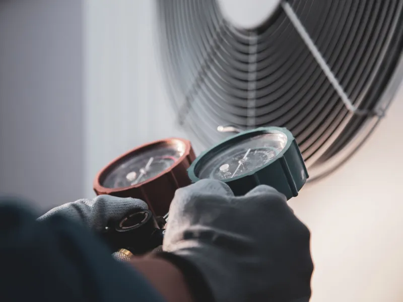 Technician wearing gloves using HVAC manifold gauges to check air conditioning system pressures near a ventilation fan.