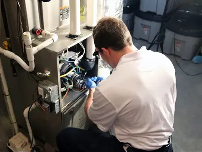 Technician in white shirt repairing a furnace with tools in a basement setting with pipes and storage bins nearby