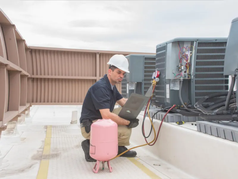 HVAC technician wearing hard hat inspecting rooftop air conditioning units with laptop and tools