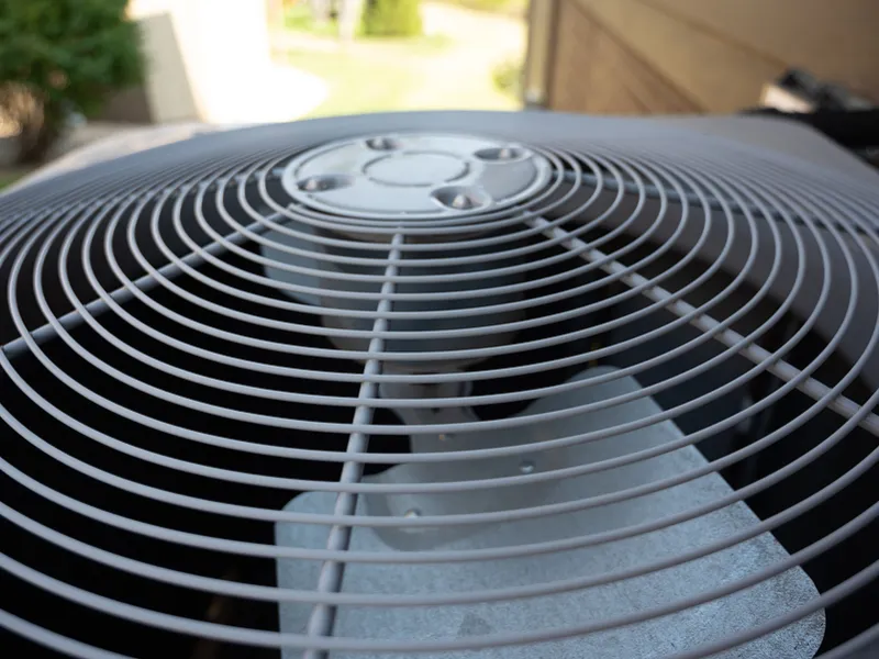 Close-up of an outdoor air conditioning unit fan with protective grille on a sunny day.