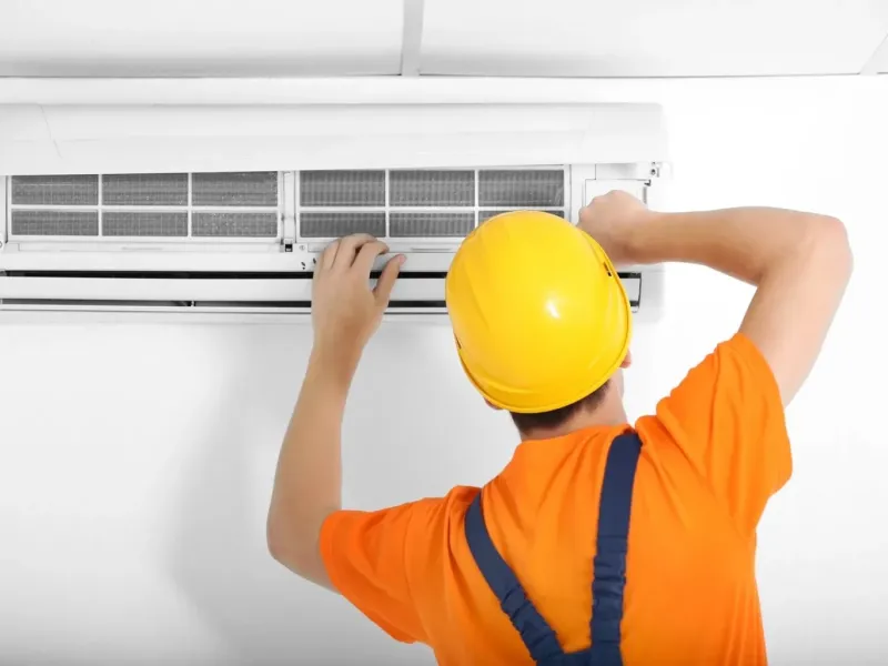 Technician wearing yellow helmet and orange shirt repairing a wall-mounted air conditioner indoors.