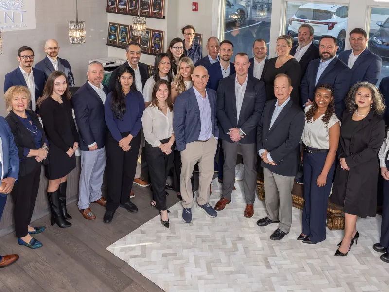 Group photo of diverse professionals in business attire inside an office with Reliant Real Estate branding.