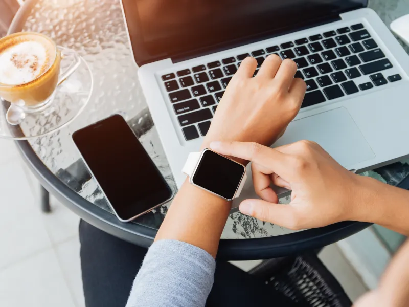 Person using smartwatch near laptop, smartphone, and coffee on glass table in bright setting