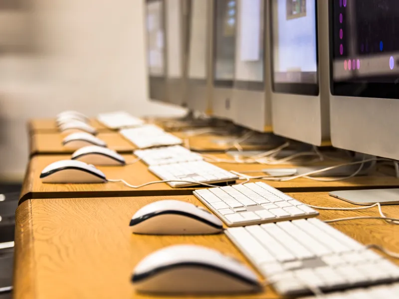 Row of desktop computers with keyboards and mice on a wooden table in a modern computer lab or office.