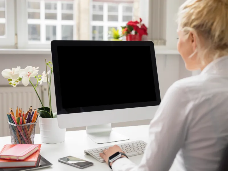 Woman typing on keyboard at desk with computer, flowers, pencils, smartphones, and notebooks near window.
