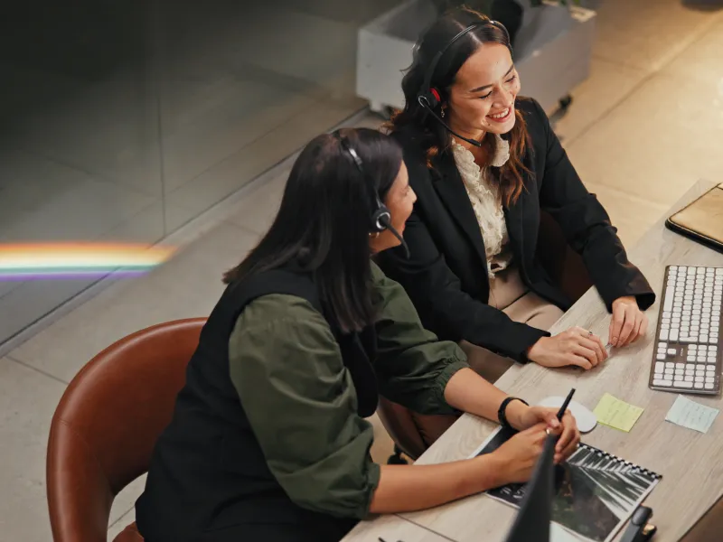Two women wearing headsets collaborate at a desk with a computer, keyboard, and notepad in a modern office.