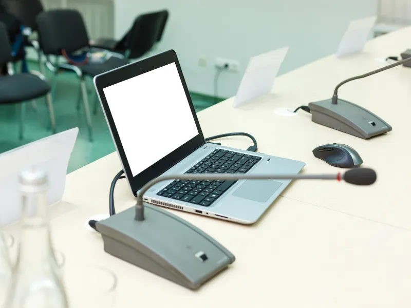 Laptop with blank screen and conference microphone on white meeting table in an empty conference room.