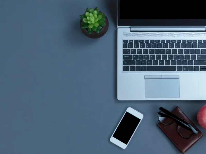 Overhead view of a laptop, smartphone, succulent plant, wallet with glasses, and apple on a gray desk surface.