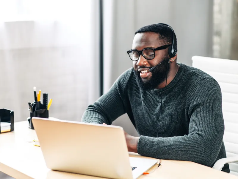 A man wearing glasses and a headset smiles while working on a laptop at a bright office desk.