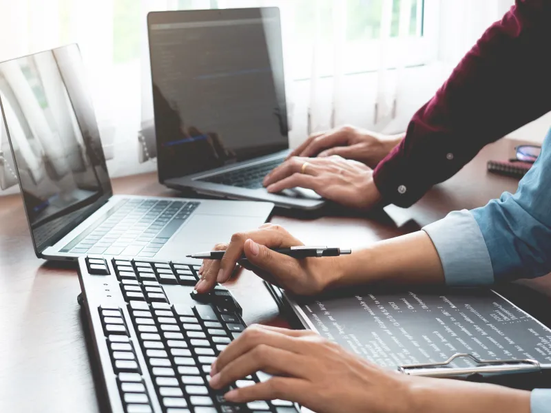 Two software developers coding on laptops in a bright office with multiple screens and wooden desk.