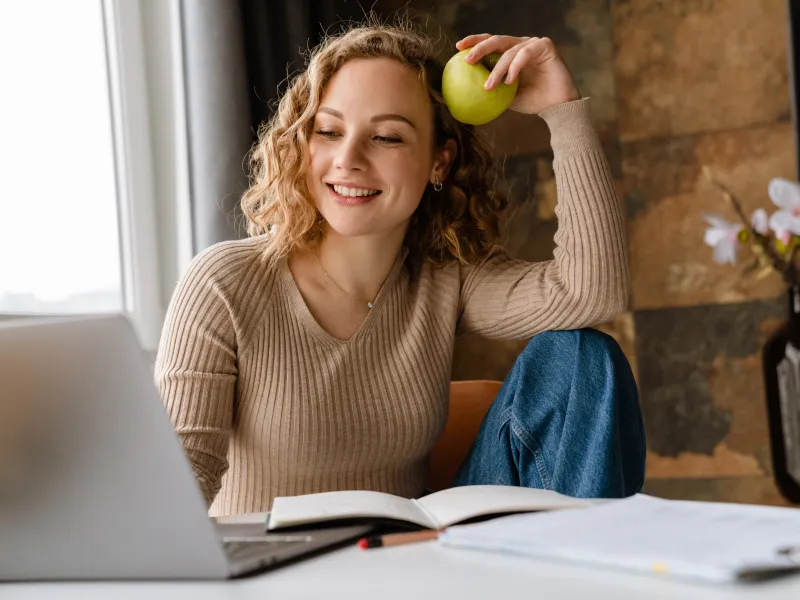 Smiling young woman holding green apple while sitting by laptop and open book in cozy room with flowers