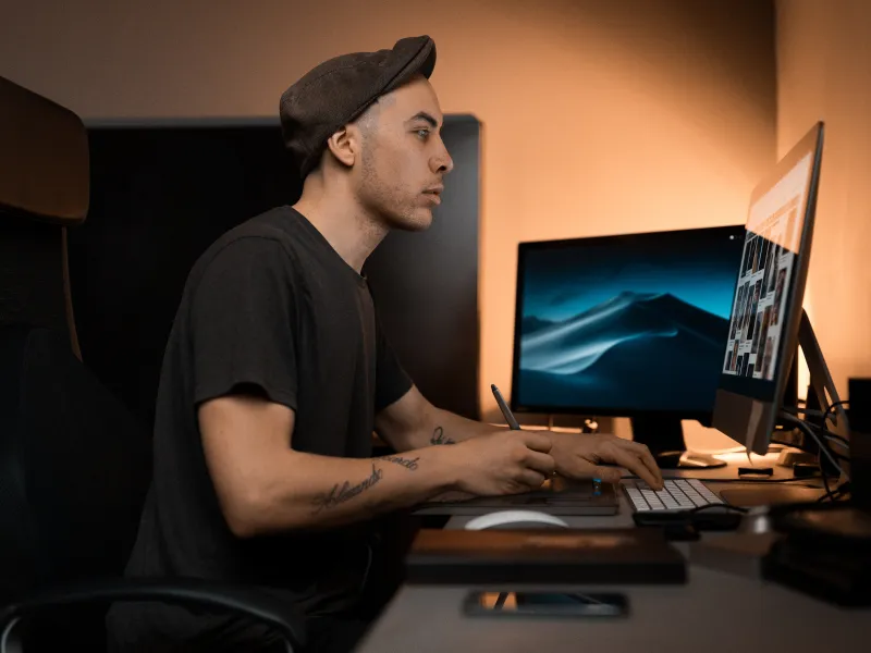 Young man wearing a cap working on digital art at a computer desk with dual monitors in a dimly lit room.