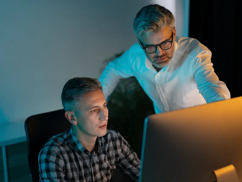 Two men working late, focused on a desktop computer screen in a dimly lit office setting.