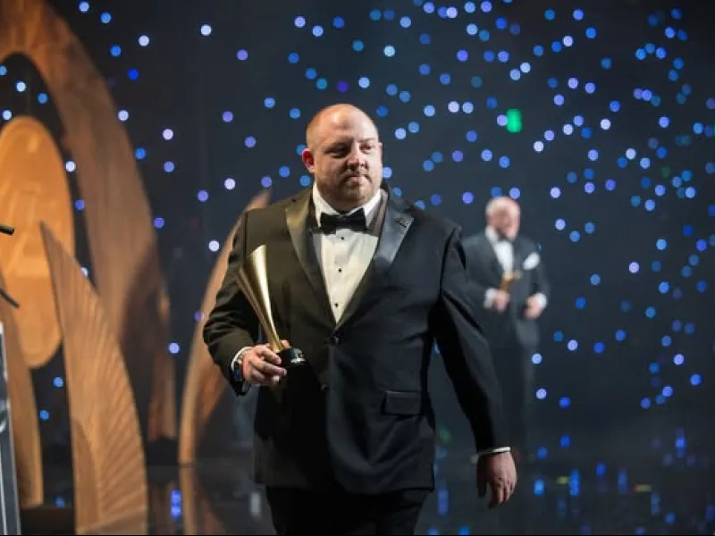 Man in tuxedo holding golden award on stage with decorative lights and large backdrop elements.