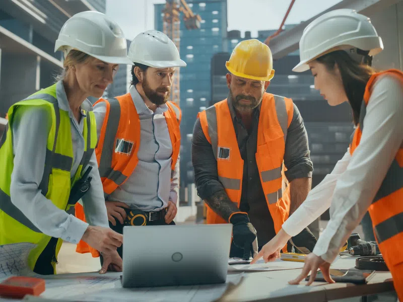 Four construction workers wearing helmets and safety vests reviewing plans and laptop at a building site.