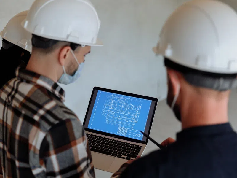 Construction workers wearing helmets reviewing a building blueprint on a laptop at a construction site.