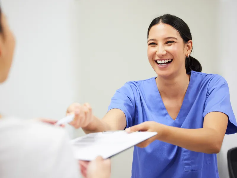 Smiling healthcare professional in blue scrubs handing a clipboard to a patient in a medical setting.