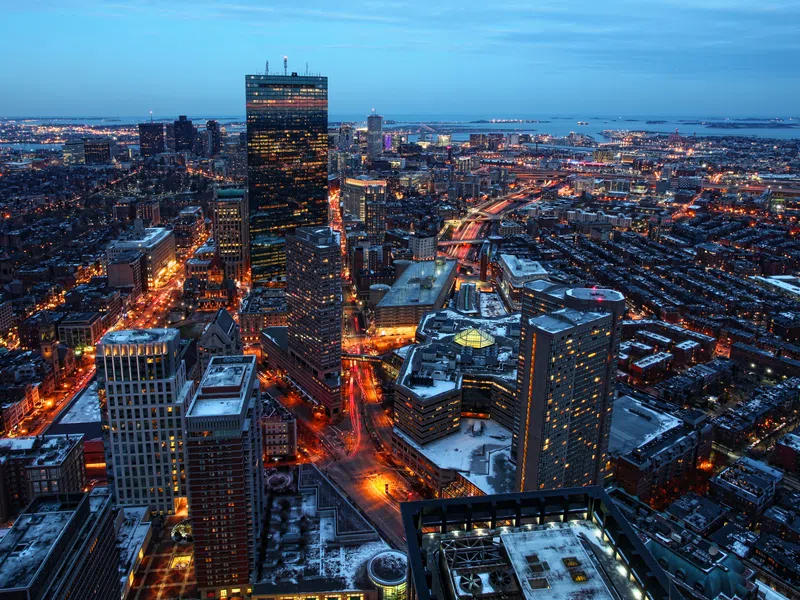 Aerial view of a city skyline at dusk with illuminated buildings, streets, and distant waterfront under a blue sky.