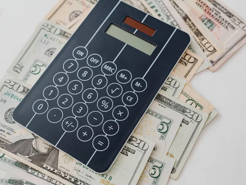 Black calculator on stacked US dollar bills of various denominations on a white surface.