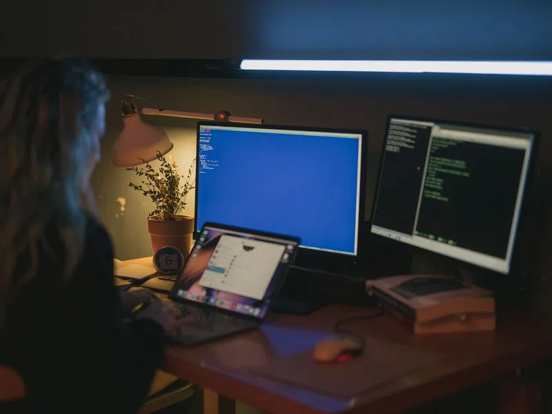 Person working on a laptop with two monitors displaying code in a dimly lit workspace with plants and a desk lamp.