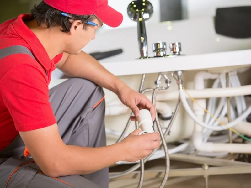 Plumber in red shirt and cap repairing white PVC pipes under a sink with exposed plumbing and faucet.