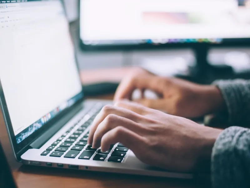 Hands typing on a laptop keyboard with blurred monitors in the background in a work setting.