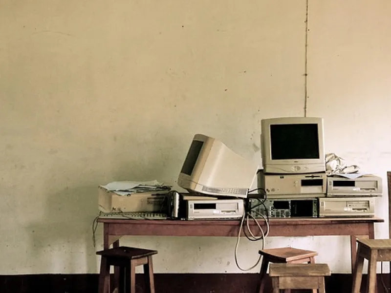 Old vintage computer monitors and desktop units stacked on a wooden table with stools in a sparse room.