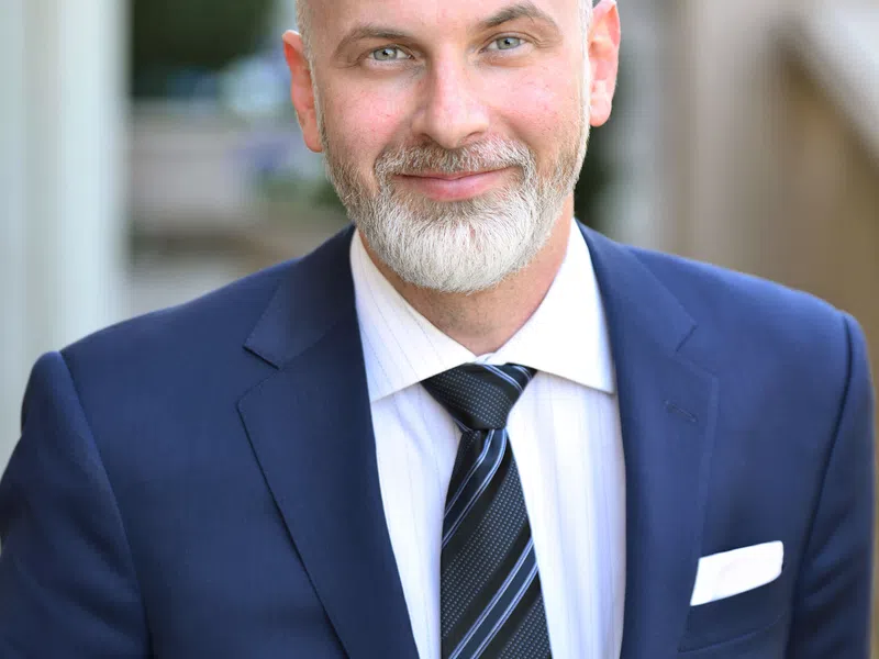 Smiling mature man with gray hair and beard wearing blue suit and striped tie in outdoor professional headshot.