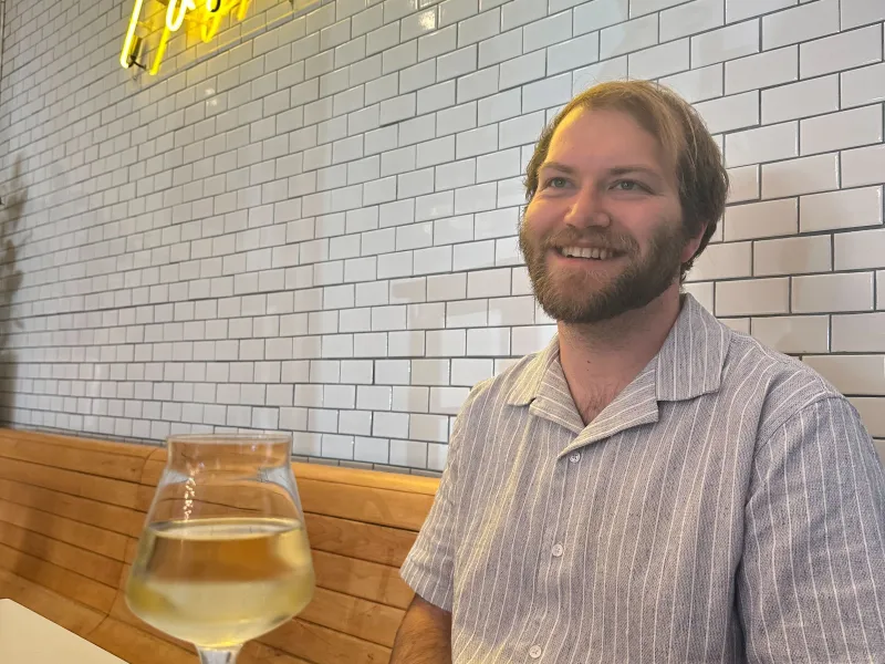 Man smiling while sitting in a modern cafe with white tiled wall and yellow neon sign behind him.