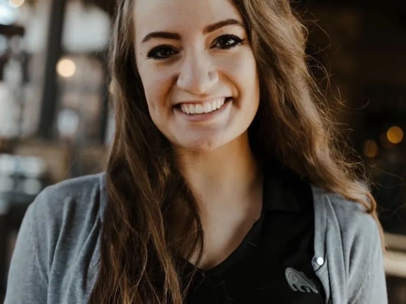 Young woman with long brown hair smiles warmly wearing a black shirt and gray cardigan in cozy indoor setting