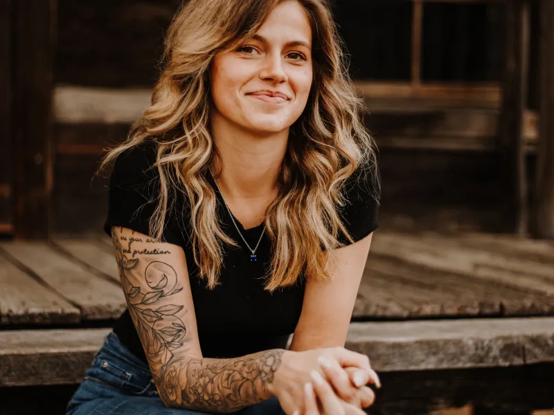 Young woman with wavy blonde hair and tattoos on arm sitting on wooden steps outside rustic building