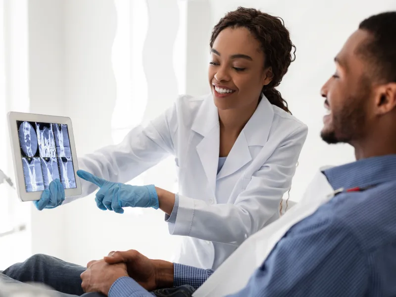 Dentist explaining dental X-ray results to smiling male patient in clinic using tablet screen